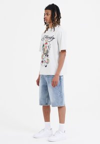 Camiseta gráfica de color gris claro con un diseño impreso colorido, combinada con pantalones cortos de mezclilla azul claro. El modelo lleva zapatillas blancas y calcetas cortas.