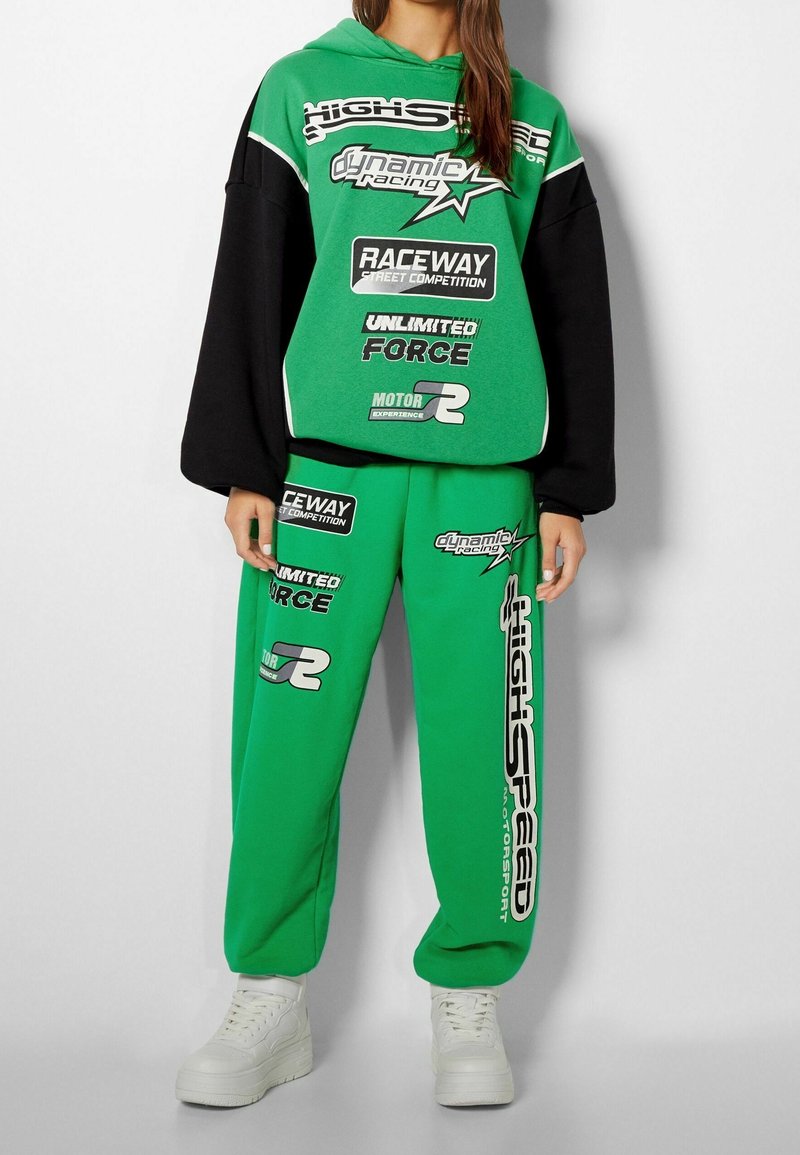 Grüne Kapuzenjacke mit schwarzen Ärmeln und weißen Akzenten, ausgestattet mit rennthematischen Grafiken. Passende grüne Jogginghosen mit auffälligen Logos. Weiße Turnschuhe.