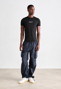 Man wearing black Calvin Klein t-shirt, navy cargo pants, and white sneakers standing on a light floor against a plain white wall.