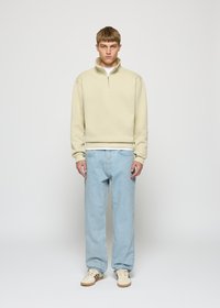 Ung mann står, iført beige kort glidelås-genser, lyseblå jeans og hvite joggesko med brune striper, foran en ensfarget hvit bakgrunn.