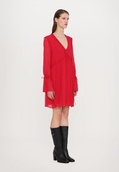 Robe rouge à manches longues avec des bords volants, décolleté en V et accents à nouer aux manches. Portée avec des bottes noires à talons hauts.