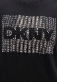 Μαύρο ύφασμα με μεγάλες γράμματα "DKNY" περιγραμματισμένα από ένα πυκνό μοτίβο μικρών, γυαλιστερών ασημένιων καρφίτσων που σχηματίζουν ένα ορθογώνιο σχέδιο.