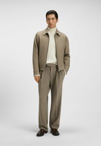 Giacca beige con colletto, dolcevita in maglia a trecce crema e pantaloni a gamba larga coordinati. Scarpe marrone scuro completano l'outfit.