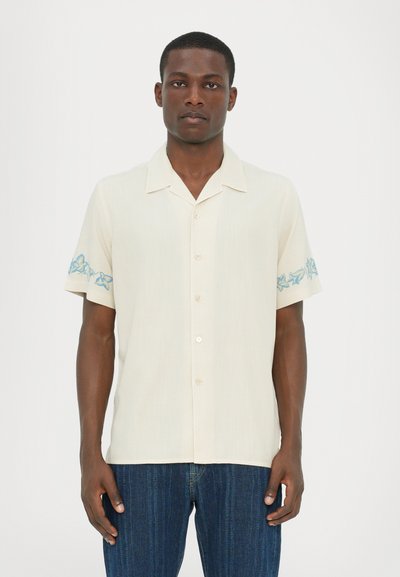 Camisa de manga corta color crema con botones y bordado floral azul en las mangas. Confeccionada en tela ligera, cuenta con cuello campestre y un corte relajado.