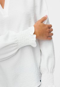 La mano di una persona è appoggiata su un braccio che indossa una blouse bianca con una texture, con polsini elastici e arricciati e una scollatura a V.
