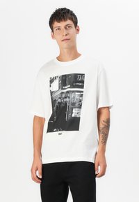 White cotton t-shirt featuring a large black-and-white photo print of a bus scene. Short sleeves and relaxed fit. Minimal branding at the hem.