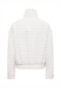 Veste blanche à manches longues à pois noirs, col montant, taille élastique et poignets boutonnés, vue de dos sur fond blanc.