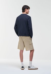 Hombre de pie de espaldas, vestido con sudadera azul marino, pantalones cortos cargo beige, calcetas blancas y zapatillas blancas con suelas marrones.