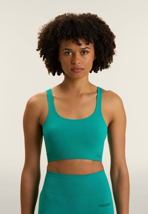 Donna con capelli ricci che indossa un reggiseno sportivo turchese e leggings a vita alta, in piedi contro uno sfondo chiaro e semplice.