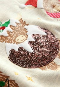 Ornement marron en sequins avec un visage souriant, des accents blancs et un chapeau de Père Noël rouge sur un fond de tissu crème clair avec des étoiles dorées.