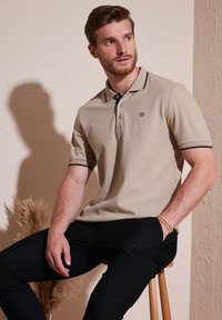 Beige pólóing fekete részletekkel és gombokkal, rövid ujjú, fekete nadrággal viselve. A modell egy fa széken ül egy textúrázott fal előtt.
