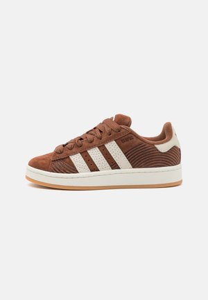 Brown suede sneaker with white leather accents, three stripes design, textured side panels, and a rubber sole. Features a lace-up closure.