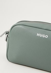Grønn skinnpose med teksturert overflate, med en sølvglitrende glidelås og hvit "HUGO"-merking på fronten. Kompakt og rektangulær form.