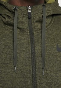 Sweat à capuche zippé vert foncé en tissu texturé. Comprend des cordons de serrage, une fermeture éclair noire et un petit logo noir sur la poitrine.