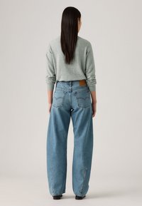 Jean en denim bleu clair à taille haute avec des jambes larges, présentant une étiquette en cuir au dos et des poches arrière traditionnelles. Texture lisse, design décontracté.