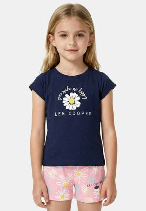 Jeune fille portant un t-shirt bleu marine avec une marguerite et le texte « tu me rends heureux », associé à un short rose orné de motifs de marguerites blanches.