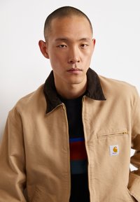 Jachetă din pânză bej cu guler din fetru maro, buzunar frontal cu fermoar și patch cu logo Carhartt. Purtați peste o cămașă neagră cu dungi colorate.
