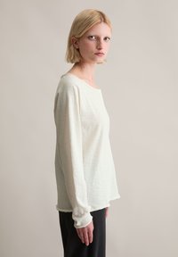 Leicht strukturiertes, off-white Langarmshirt mit rundem Ausschnitt und lockerer Passform, kombiniert mit schwarzen Hosen, seitlich betrachtet.