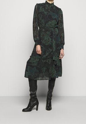 Kobieta ubrana w czarną sukienkę midi w zielony kwiatowy wzór, z długimi rękawami, falbanami na dole i wiązaniem w talii, w połączeniu z czarnymi, wysokimi kozaki na obcasie.