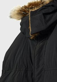 Manteau d'hiver noir avec capuche bordée de fausse fourrure épaisse marron et beige, vu de dos et de côté sur un fond uni.