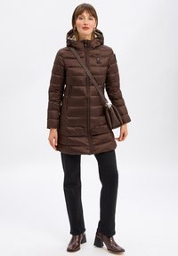 Bruine pufferjas met horizontale stiksels, een capuchon, twee frontzakken en een zwarte rits. Gecombineerd met zwarte broeken en bruine laarzen met hak.