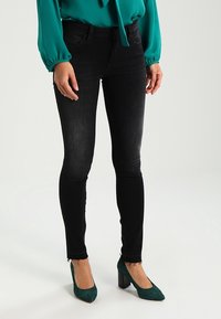 Jeans skinny negros con un lavado medio, con una cintura alta, ajustados y con dobladillo deshilachado, combinados con tacones de ante verdes.