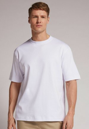 Jeune homme aux cheveux courts portant un t-shirt blanc uni à manches courtes et un pantalon beige, debout devant un fond neutre.