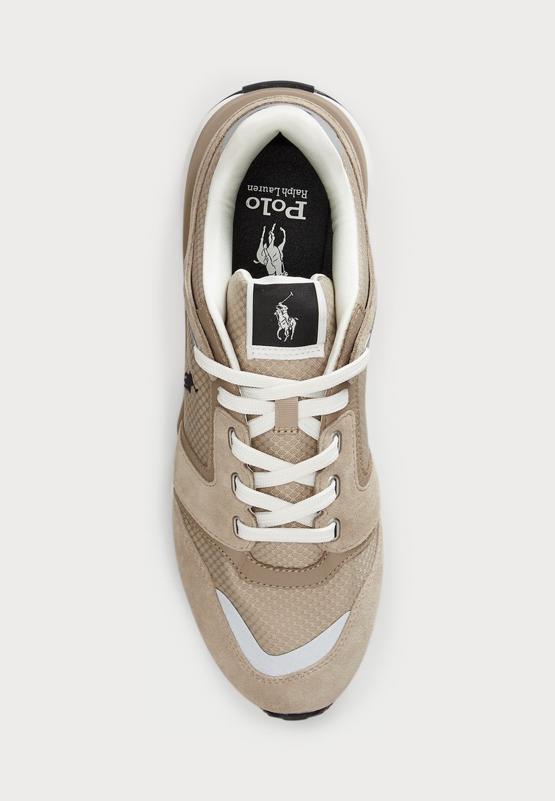 Zapatilla beige con exterior de malla y ante; cordones blancos, detalles plateados y una plantilla negra con un logo. Diseño de caña baja.