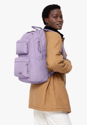 Un sac à dos lilas avec plusieurs poches avant, des zips et des bretelles rembourrées, porté sur un manteau marron, sur fond blanc uni.