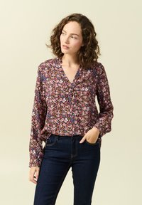 Långärmad blommig blus med djup v-ringning, med en mörk bakgrund och flerfärgade blommönster. Bärs med mörka jeans.