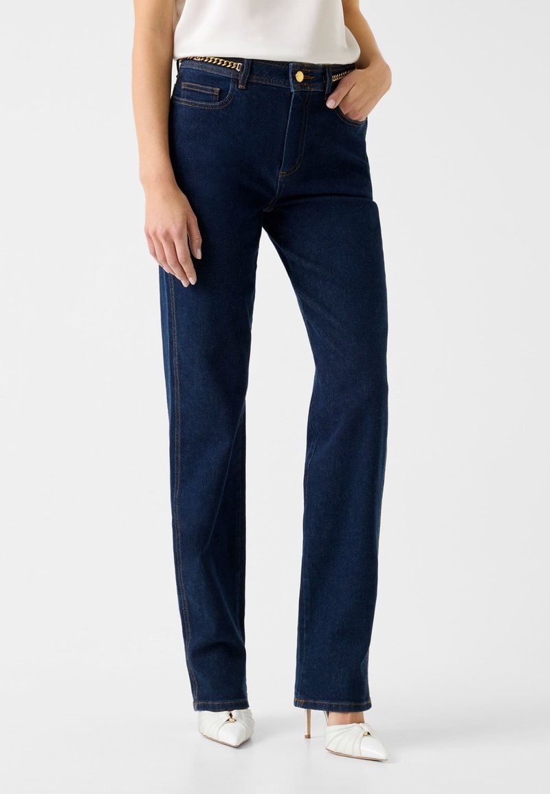 Frau trägt dunkelblaue Jeans mit geradem Bein, einem goldenen Kettengürtel und weißen spitzen High-Heels, steht vor einem schlichten Hintergrund.