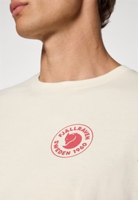 Krémové tričko s červeným kruhovým logem, na kterém je stylizovaná liška a text "FJÄLLRÄVEN SWEDEN 1960". Měkký bavlněný materiál, design s kulatým výstřihem.