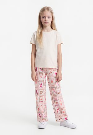 Ragazza bionda in piedi rivolta in avanti, indossa una maglietta beige chiaro semplice e pantaloni larghi con motivi rosa e marroni.