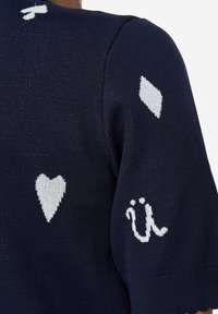 Tmavě modrý pletený svetr s bílými vzory srdcí a tvarů, s akcentem 'ü' na rukávu. Texturovaná látka s krátkými rukávy.