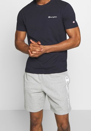 Homme portant un t-shirt noir Champion et un short gris clair Champion, debout les mains jointes devant.