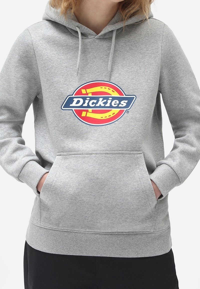 Personne portant un sweat à capuche gris avec un logo Dickies rouge, jaune et bleu centré sur la poitrine, mains dans la poche avant.
