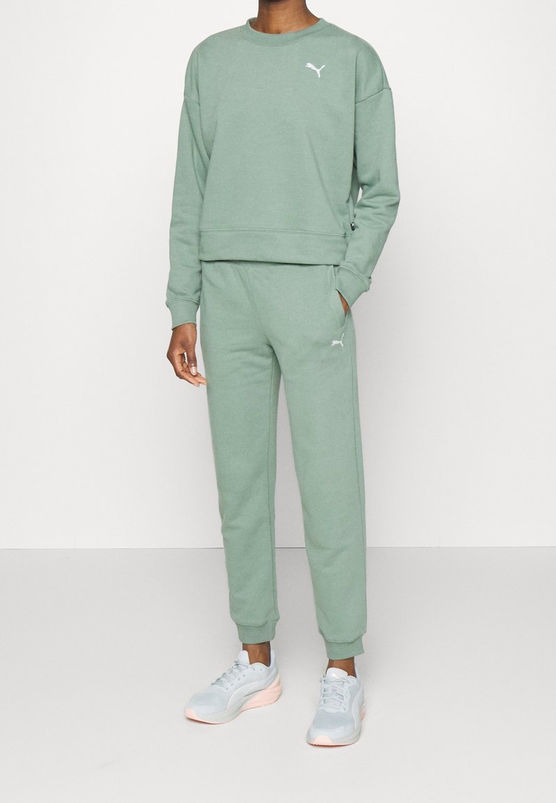Personne portant un sweat-shirt Puma vert clair et un jogging assorti avec des logos Puma blancs, les mains dans les poches, assorti à des baskets gris clair.