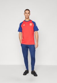 Punane ja sinine spordiriietus, mis sisaldab lühikeste varrukatega särki, millel on vapp ja valged triibud, koos tumesiniste pükste ja mustade kingadega.