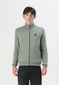 Light green zip-up jacket with a high collar, side pockets, and a logo patch on the chest. Made from a soft, smooth fabric.