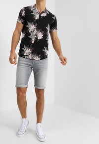 Camisa negra de botón de manga corta con un patrón floral en rosa y verde. Combinada con pantalones cortos de mezclilla gris claro arremangados y zapatillas blancas.