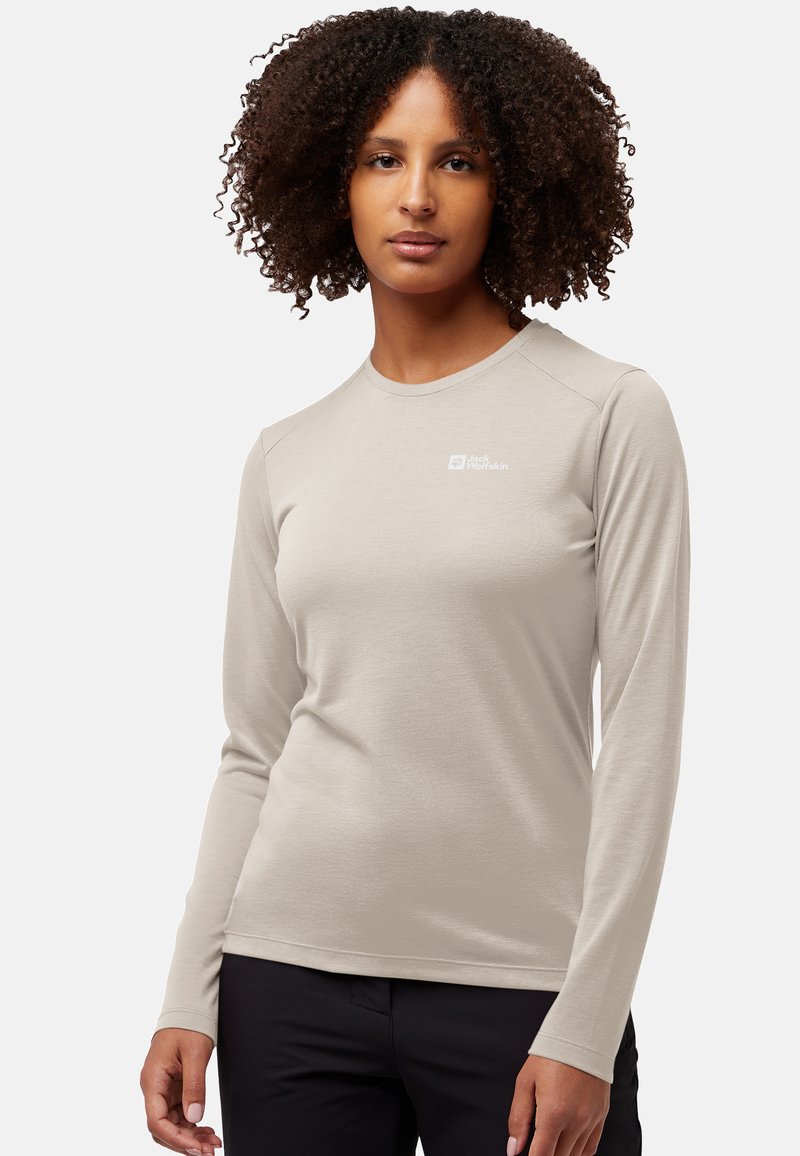 Vrouw met krullend haar, gekleed in een beige Jack Wolfskin shirt met lange mouwen en zwarte broek, staand tegen een effen lichte achtergrond.