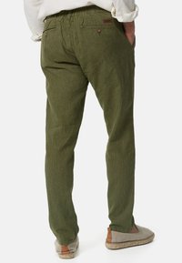 Zielone spodnie w prążki wykonane z teksturowanej tkaniny, o luźnym kroju i tylnych kieszeniach zapinanych na guziki. Noszone z jasnymi butami.