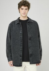 Veste chemise en denim noir avec boutons sur le devant, motif cachemire délavé, coupe décontractée et col pointu, portée avec une chemise noire en dessous.