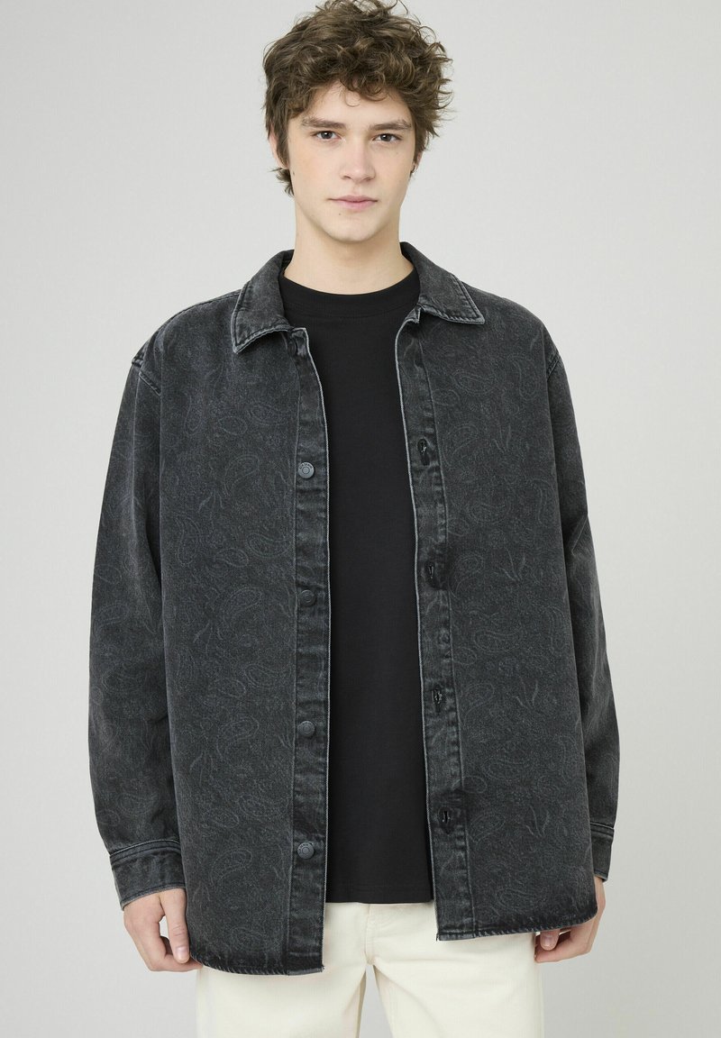 Veste chemise en denim noir avec boutons sur le devant, motif cachemire délavé, coupe décontractée et col pointu, portée avec une chemise noire en dessous.