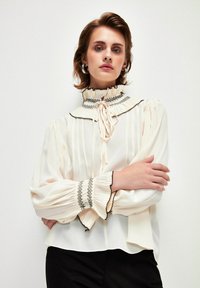 Helle cremefarbene Bluse mit einem hohen, gerafften Kragen, vertikalen Streifen und schwarzen Stickereien an den Bündchen und dem Kragen. Lange Puffärmel.