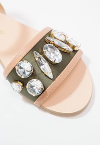 Sandale à bout ouvert avec semelle et bride beige, décorée de grandes pierres précieuses en cristal clair sur une bande vert foncé.