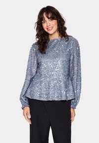Blå pailletbluse med lange puffærmer, rund halsudskæring og tekstureret stof. Løs pasform med peplum-nederdel. Parret med sorte bukser.