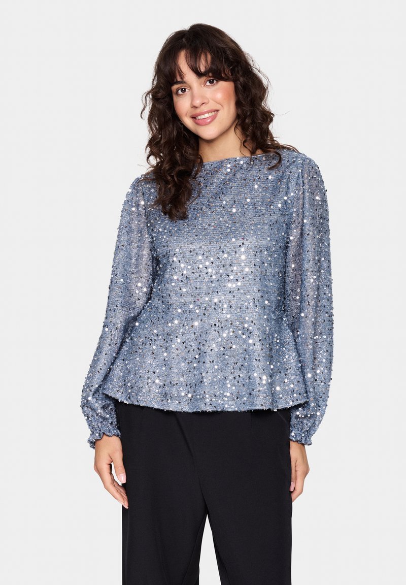 Blå pailletbluse med lange puffærmer, rund halsudskæring og tekstureret stof. Løs pasform med peplum-nederdel. Parret med sorte bukser.