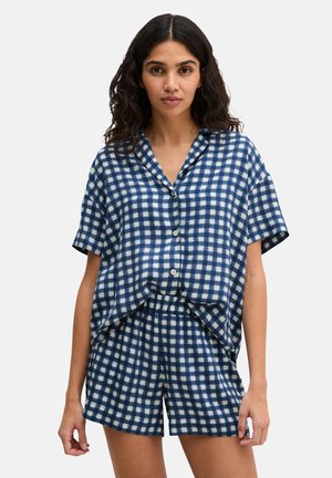 Vrouw draagt een losse blauw-wit geruite korte mouwen blouse met knopen en bijpassende short met elastische taille, staand tegen een effen achtergrond.