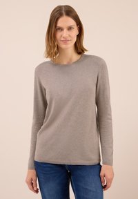 Žena s vlasy světle hnědé barvy po ramena, oblečená v jednoduchém světle hnědém svršku s dlouhým rukávem a modrých džínách, na béžovém pozadí.